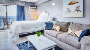 A hotel room with a bed, a sofa, a coffee table, and a painting of beach chairs. - Oceanfront Efficiency T507 (Myrtle Beach)
