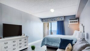 A modern hotel room with a bed, sofa, TV, dresser, and a view of the outdoors. - Oceanfront Efficiency T507 (Myrtle Beach)