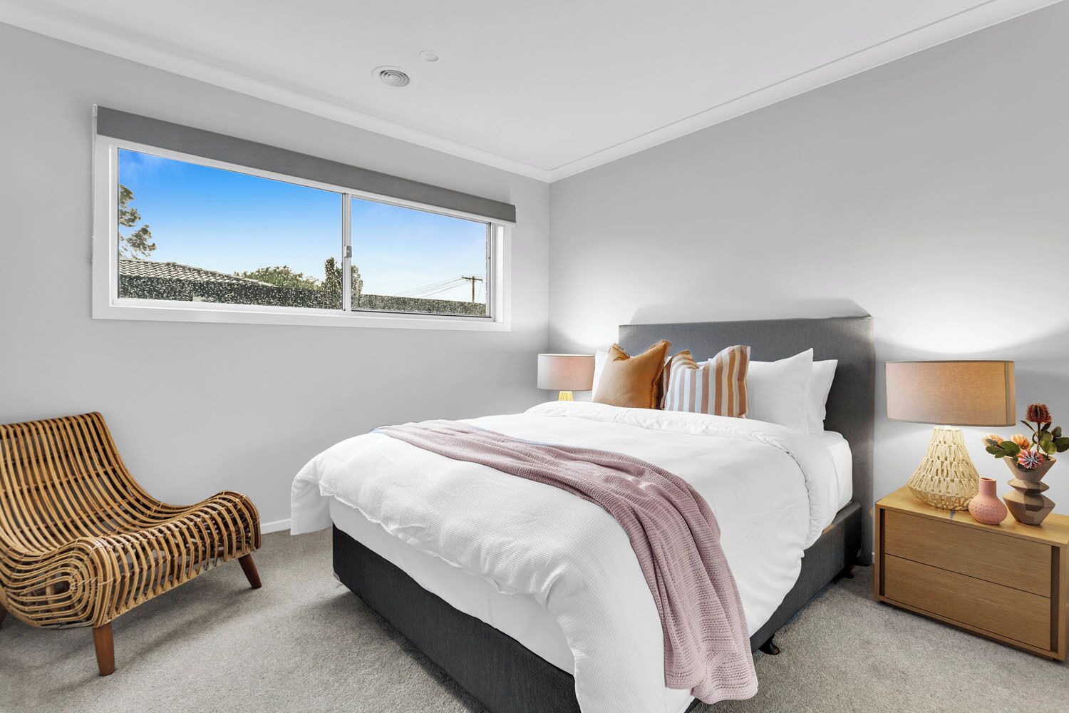 A bedroom with a large bed, a wicker chair, a nightstand with a lamp, and a window with a view of trees and a building.