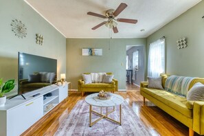 A living room with a flat-screen TV, a yellow sofa, a green armchair, and a ceiling fan.