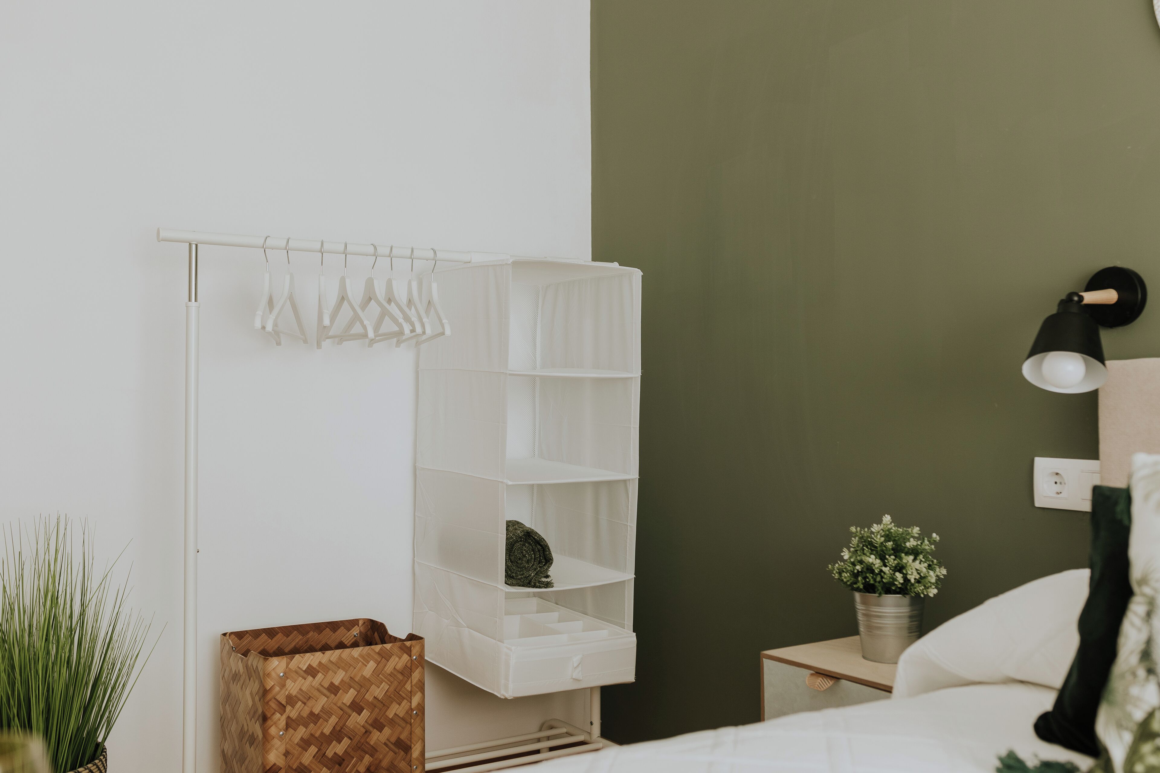 A neatly organized bedroom with a white wardrobe, a bedside table with a lamp, and a potted plant.