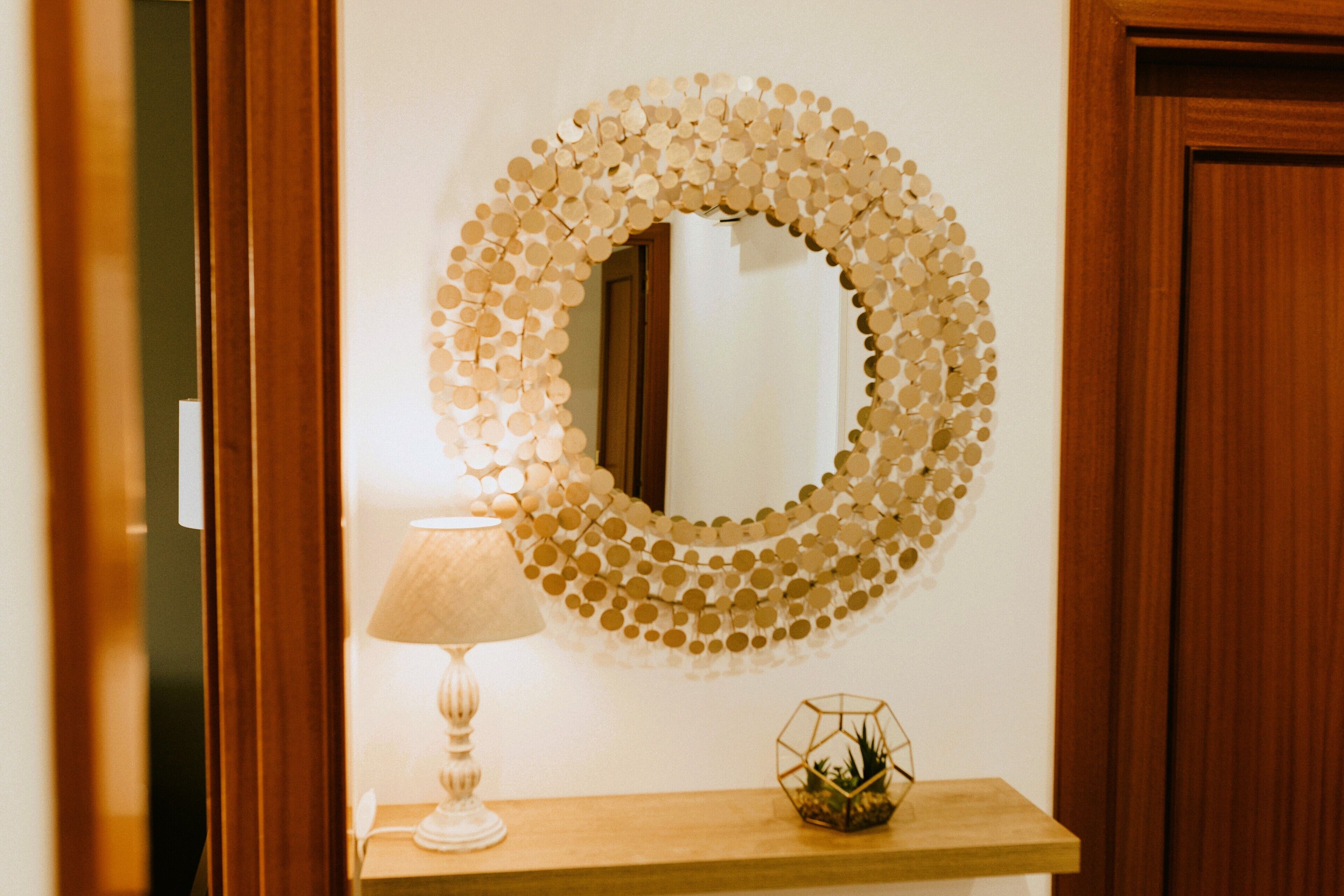 A mirror with a circular frame made of small, circular elements, a lamp, and a small decorative item on a wooden shelf.