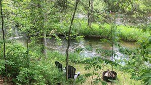 A wooden staircase leading to a seating area with chairs and a fire pit, surrounded by lush greenery and a stream.