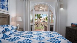 A bedroom with a bed, a nightstand, a lamp, a dresser, and a view through an arched window to a balcony with a chair.