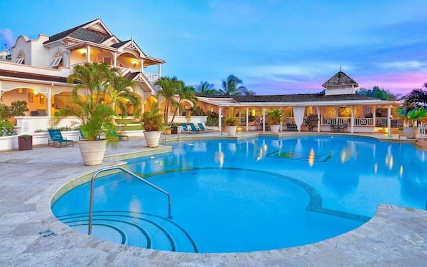 A large swimming pool with a surrounding deck, lounge chairs, and a building with multiple levels and balconies.