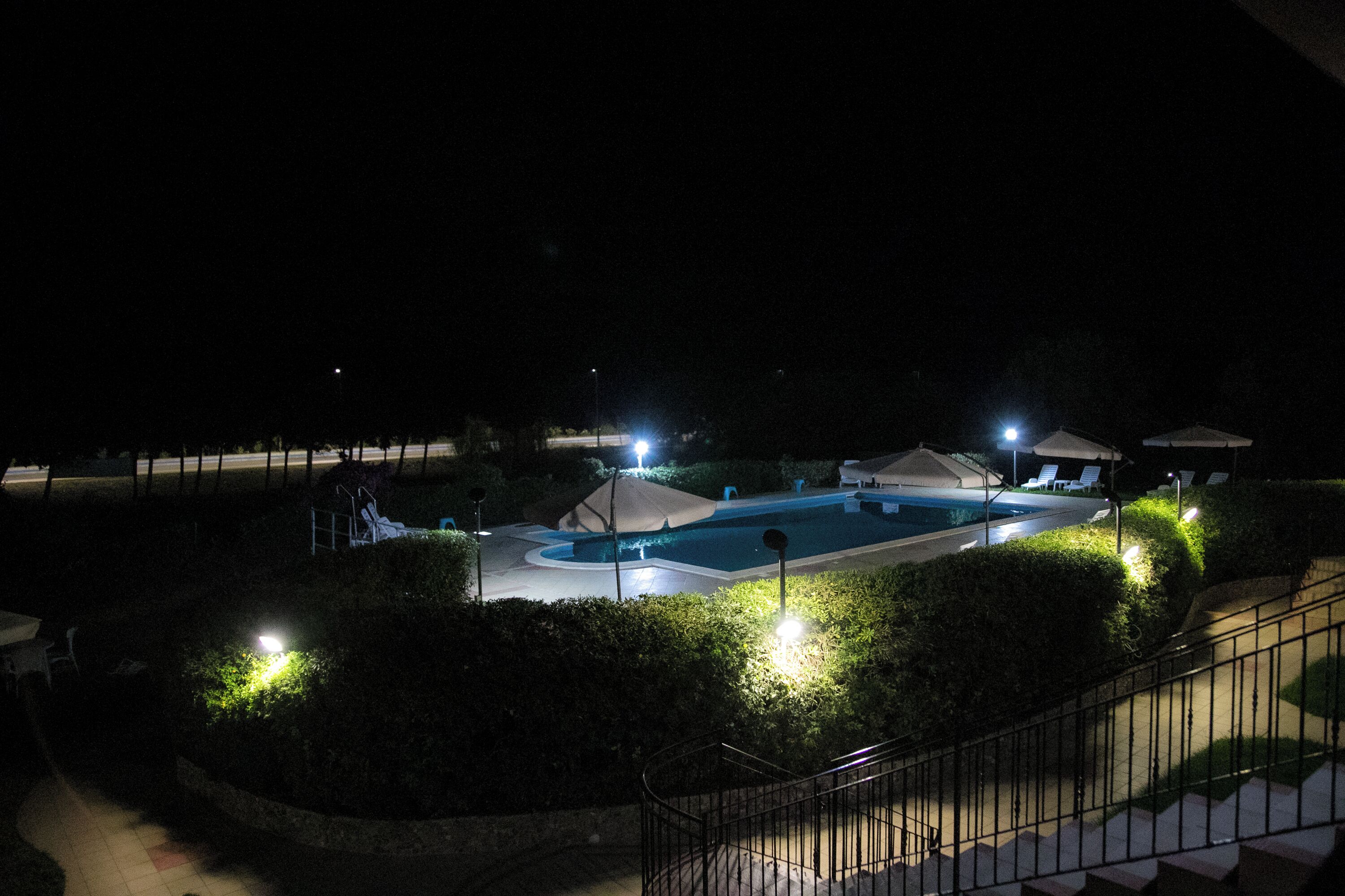 A nighttime view of a swimming pool area with surrounding greenery and a staircase leading up to a covered seating area.