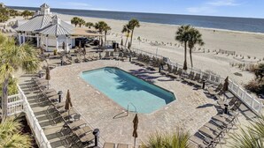 Ein Poolbereich mit Liegestühlen und Sonnenschirmen, ein Gebäude mit markantem Dach und ein Strand im Hintergrund.