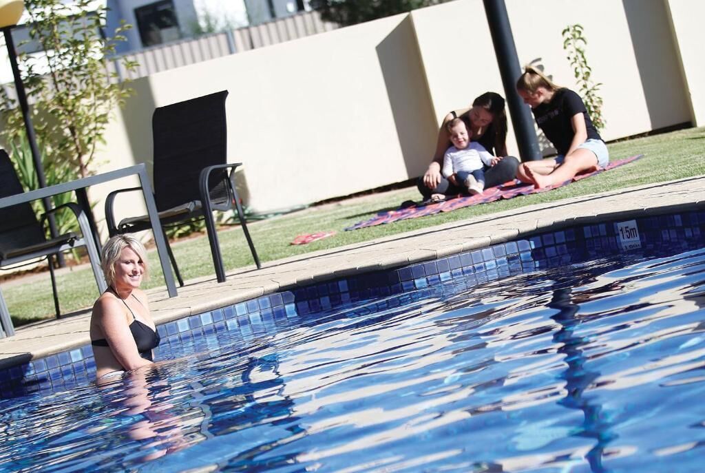 A swimming pool with a tiled edge, a grassy area with a blanket, and a building in the background.