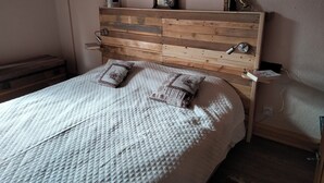 A bedroom with a wooden headboard, two pillows, a quilted bedspread, a bedside table with a lamp, and a small shelf with books and a picture frame. - Savoie, St François Longchamp, Très Grand Appartement de 180 m2, 12 Couchages (Saint-François-Longchamp)
