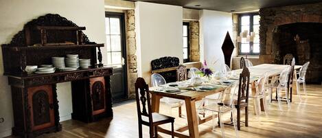 Un comedor con una mesa de madera dispuesta para cuatro personas, rodeada de sillas. A la izquierda, un aparador de madera con platos y cuencos. La habitación presenta paredes de ladrillo visto y una chimenea de piedra.