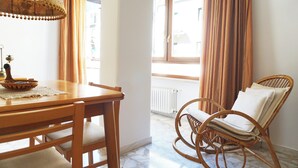 A dining area with a wooden table and chairs, a wicker chair with cushions, and a window with curtains.