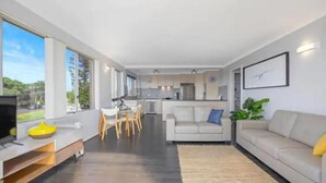 Een moderne woonkamer met een bank, een eethoek met stoelen, een keuken op de achtergrond en grote ramen met uitzicht op de omgeving.