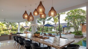 Een ruime eethoek buiten, met een lange houten tafel, zwarte stoelen en hangende rieten lampen.