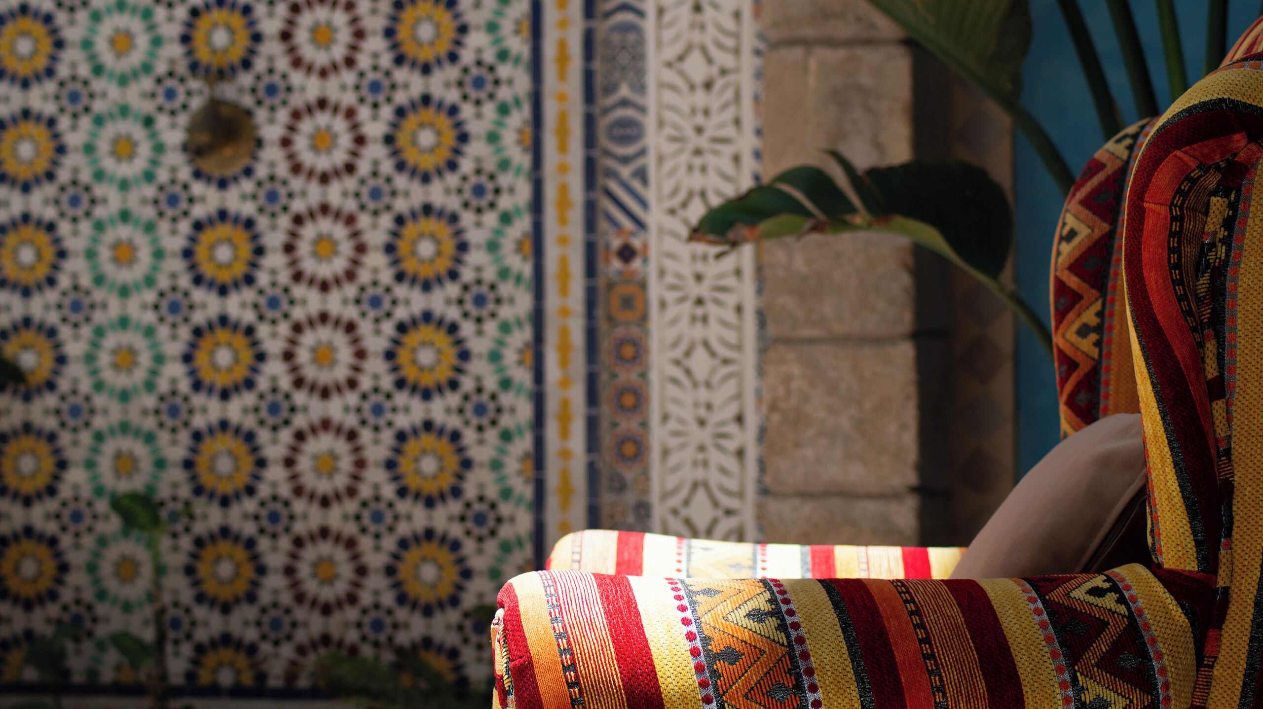 A patterned wall with a mix of geometric and floral designs, a textured fabric chair, and a potted plant.