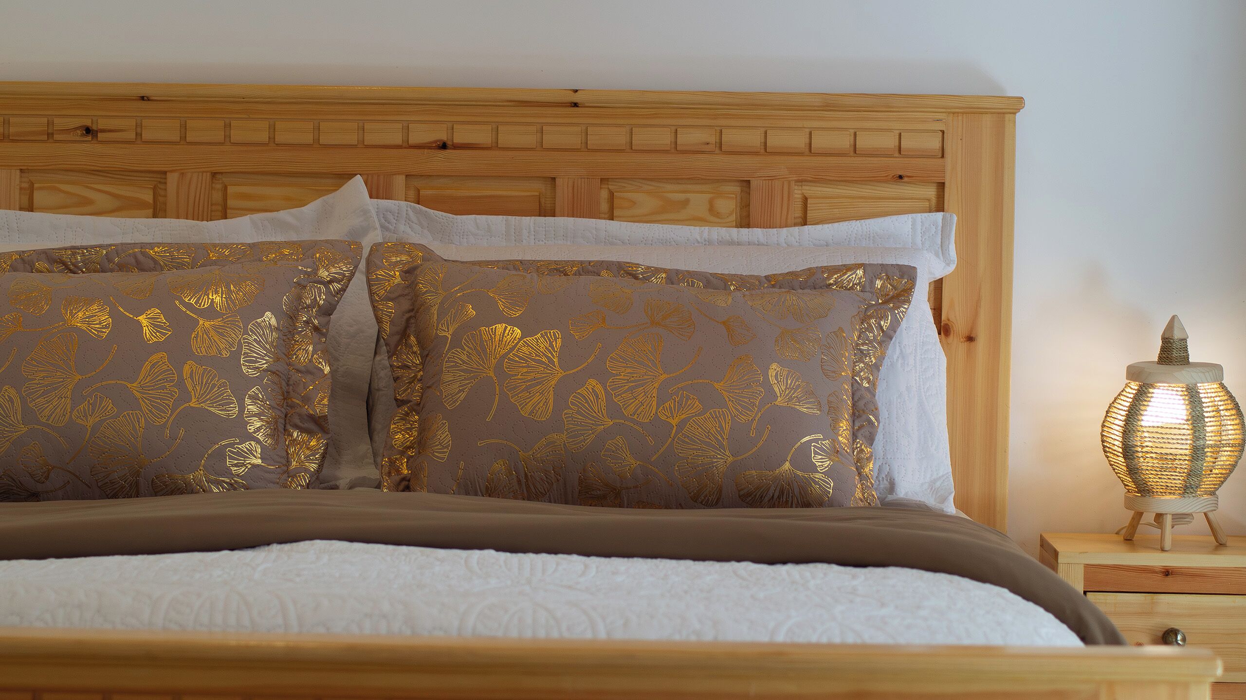 Een bed met een houten hoofdbord, twee kussens met bladmotief en een nachtkastje met een lamp.