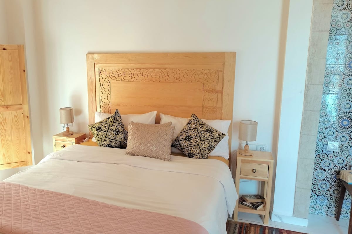 A bedroom with a large bed, two bedside tables, a wooden headboard, and a window with blue blinds.