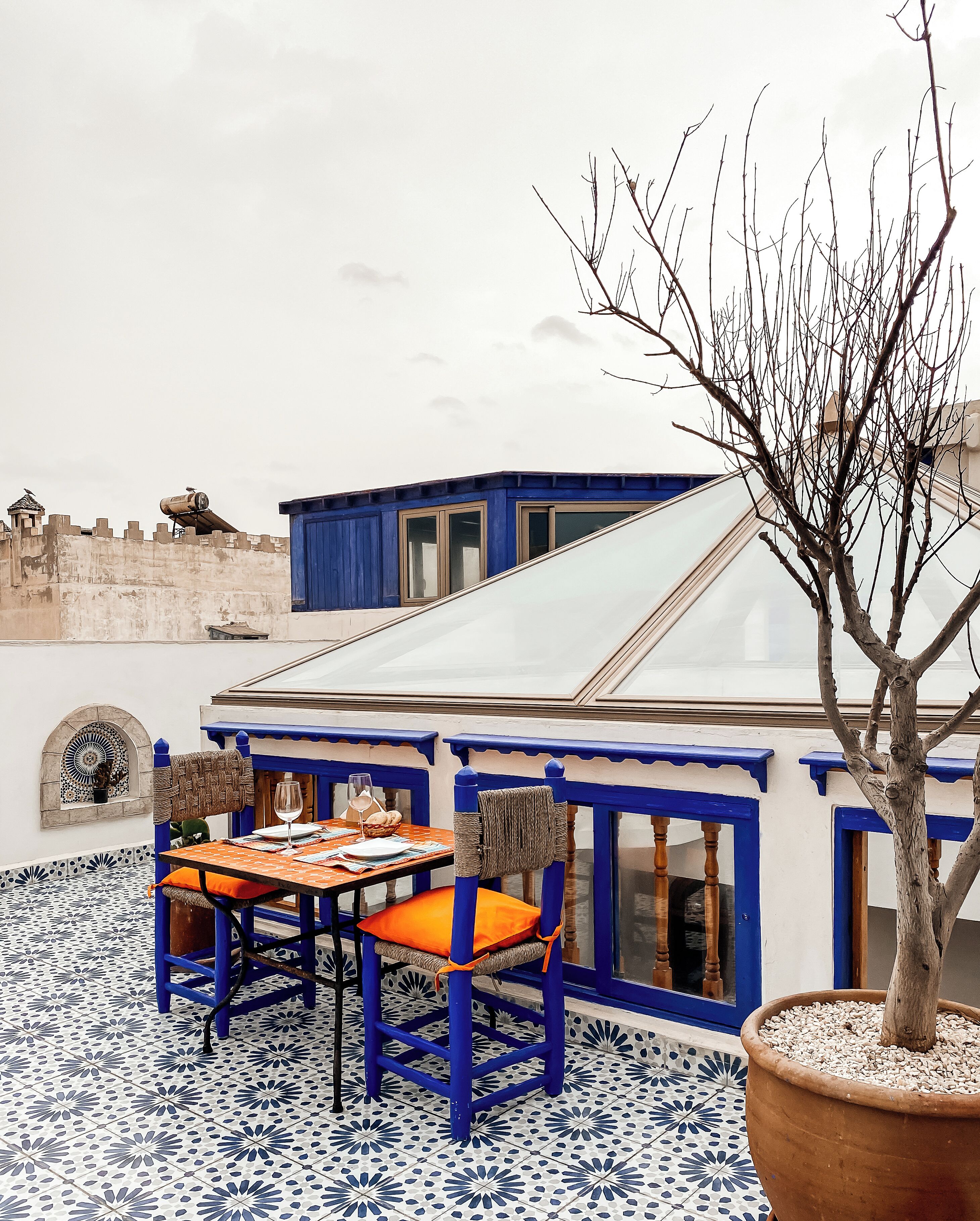 Una terraza con una mesa y sillas, un árbol en maceta y un edificio con detalles azules y un ventanal grande.