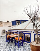 Una terraza con una mesa y sillas, un árbol en maceta y un edificio con detalles azules y un ventanal grande.