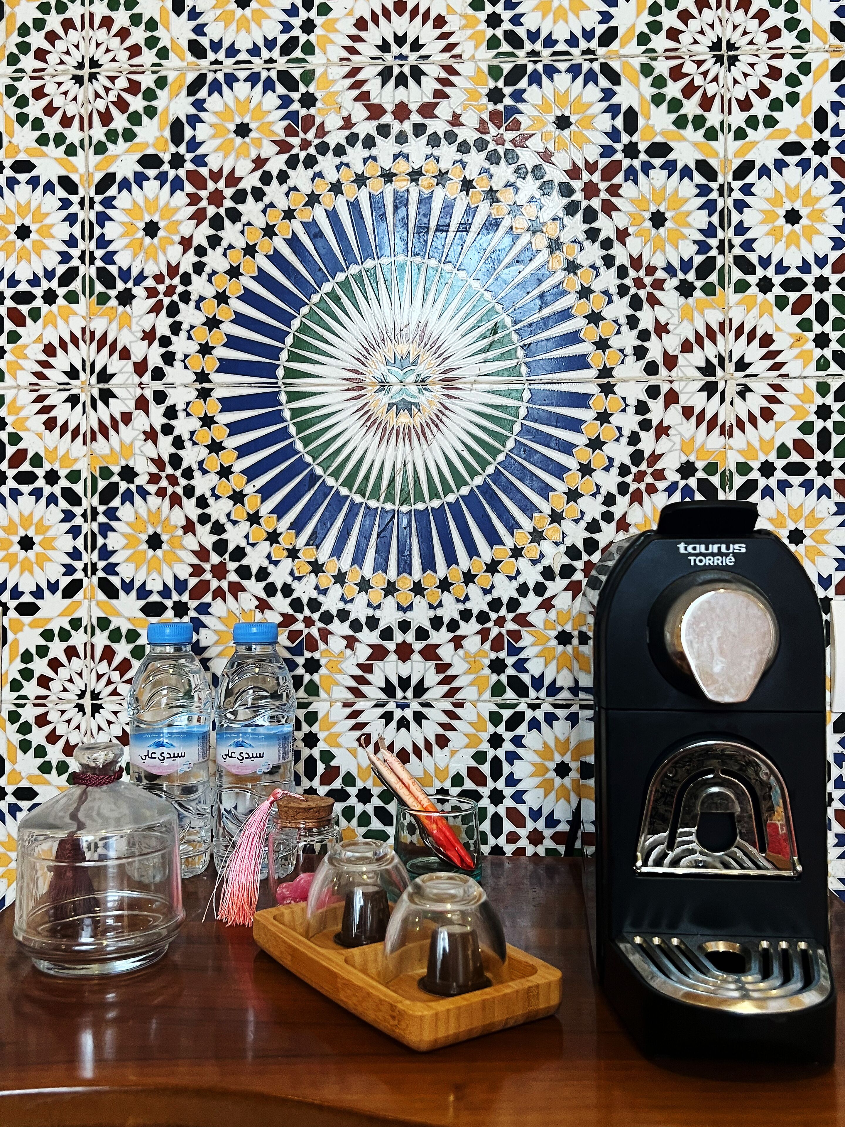 A tiled wall with a geometric pattern, a water dispenser, and various glass containers on a wooden surface.