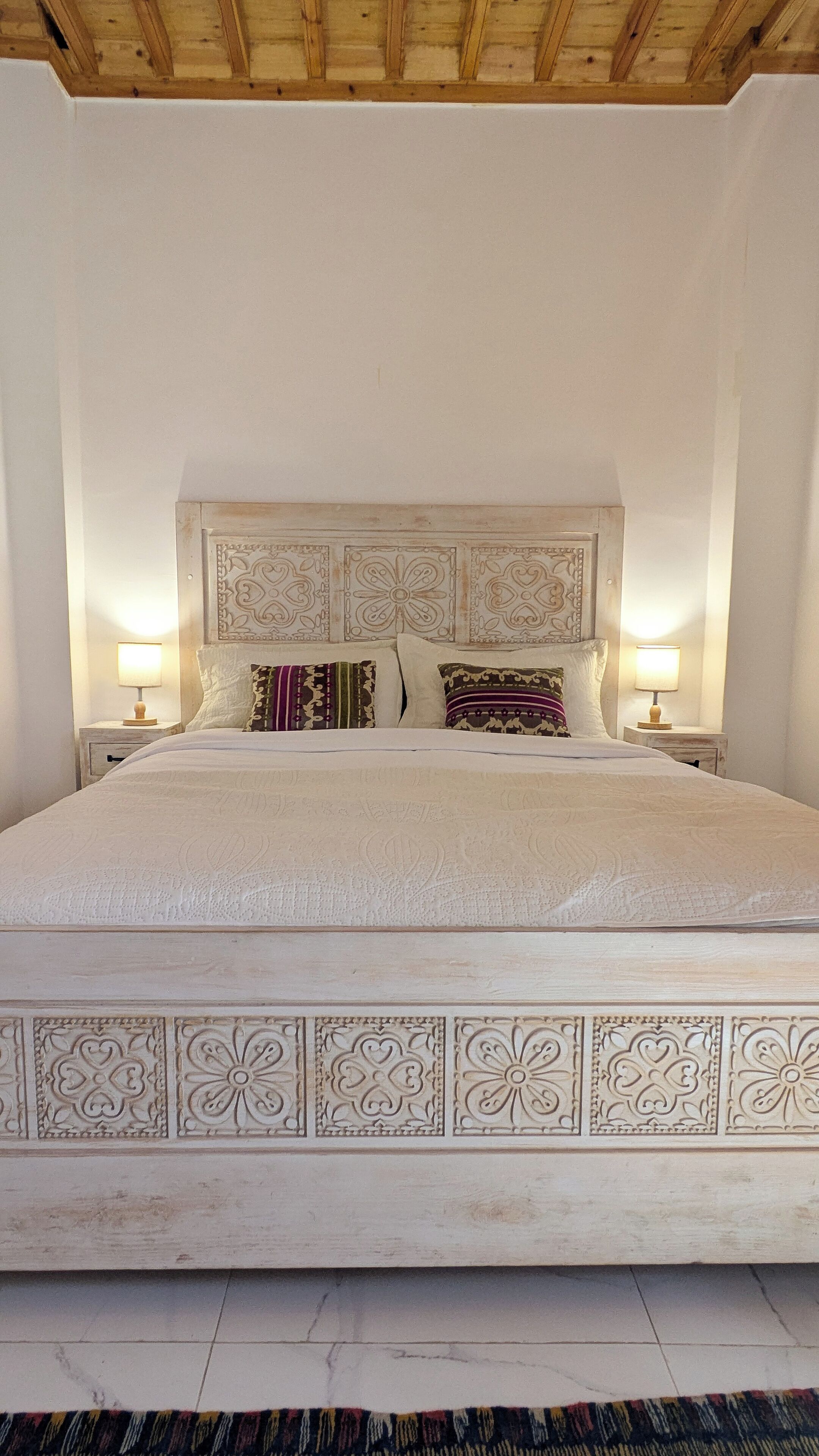 A large bed with a decorative headboard, two bedside tables with lamps, and a patterned rug on the floor.