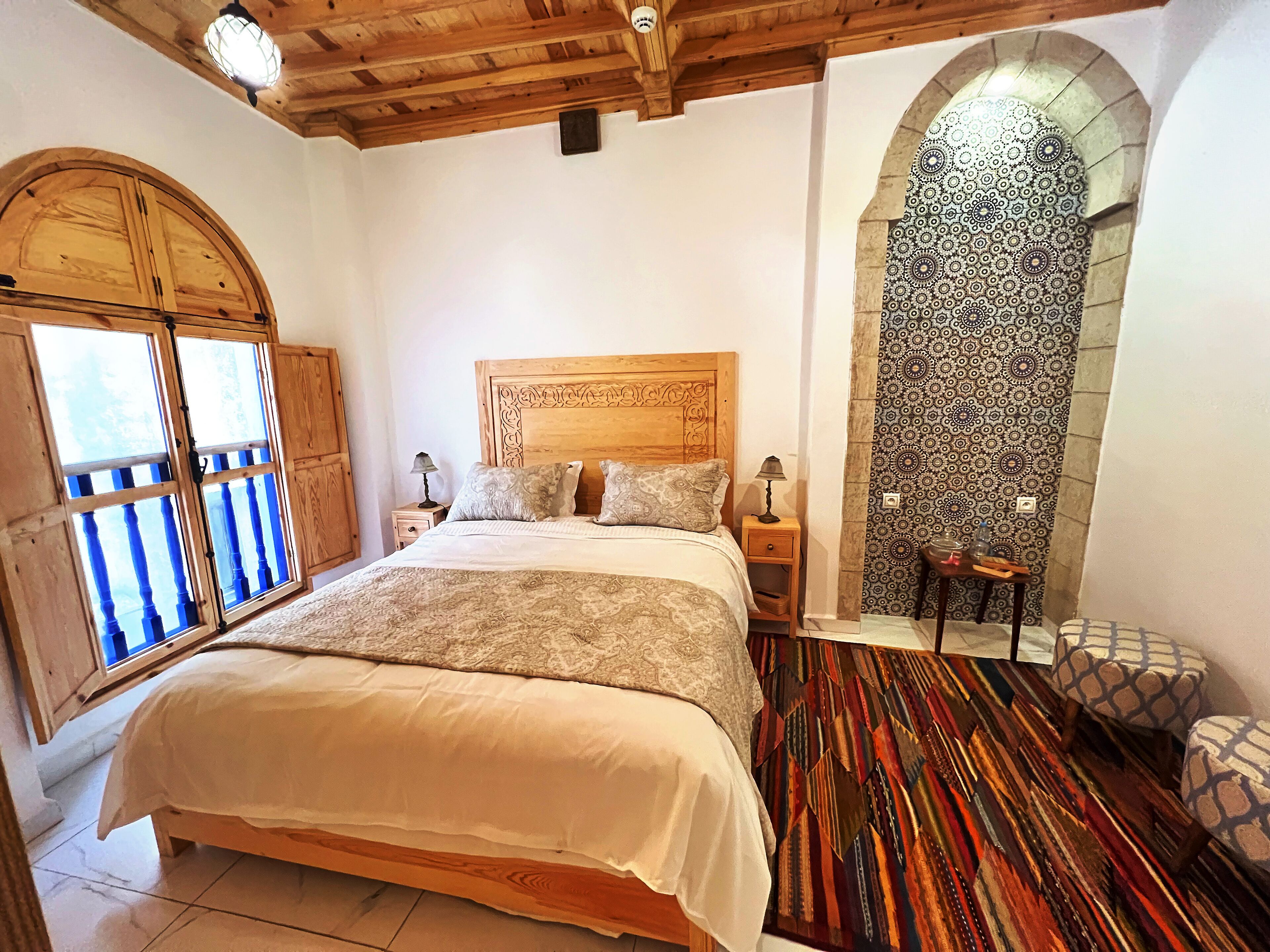 A bedroom with a large bed, wooden ceiling beams, a patterned wall, and a view through a window with a balcony.