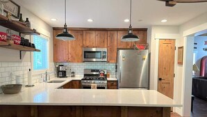 A modern kitchen with wooden cabinets, stainless steel appliances, a large island with stools, and pendant lighting. - Adventure Awaits! - Twain Harte Getaway (Twain Harte)