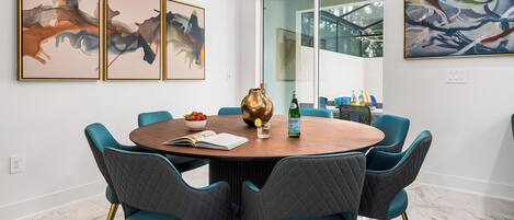 Un comedor con una mesa redonda, sillas color turquesa y patas doradas. Se observan cuadros abstractos en las paredes y una lámpara colgante moderna sobre la mesa.
