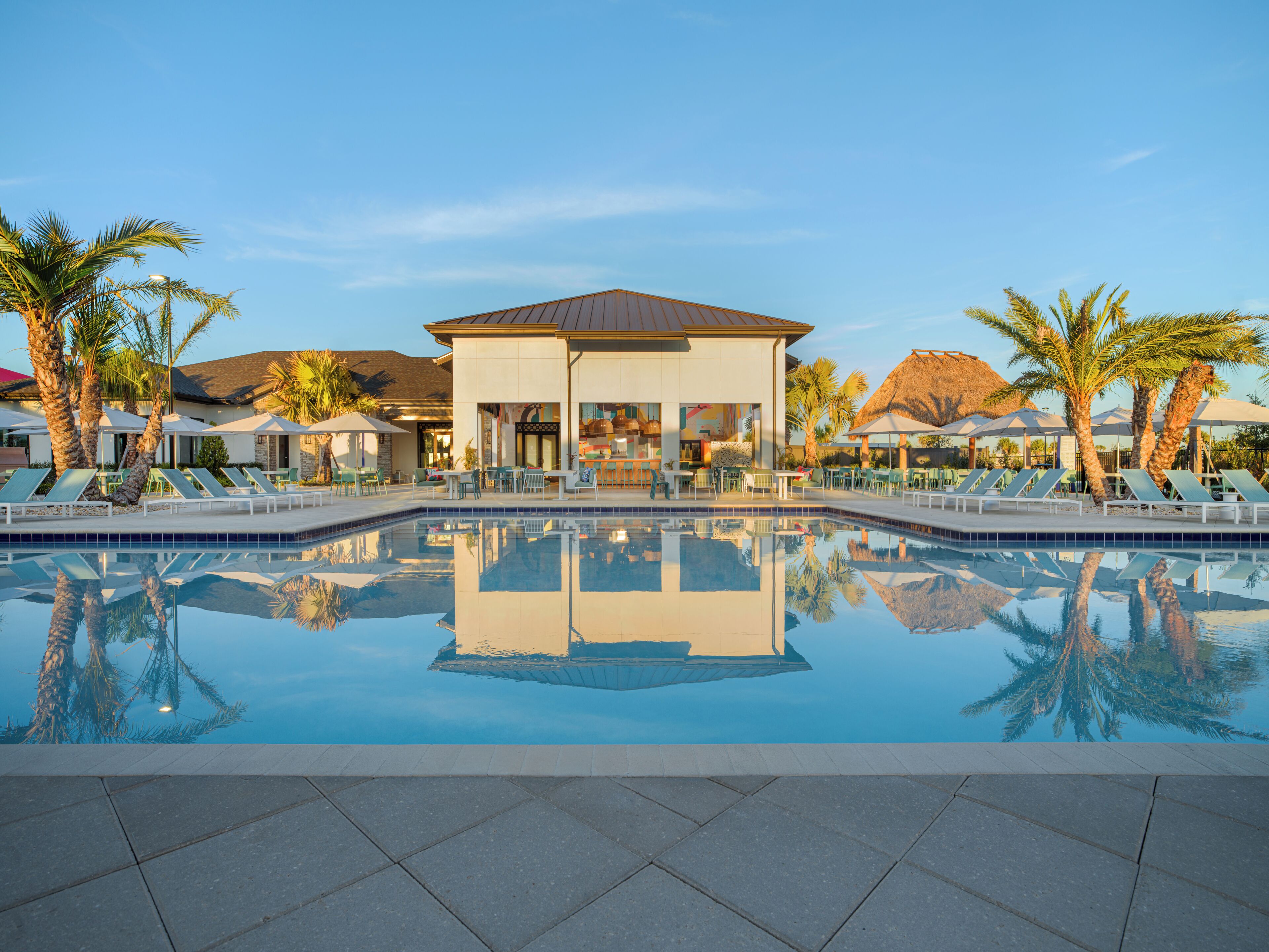 A pool area with lounge chairs and umbrellas, a building with a thatched roof, and palm trees.