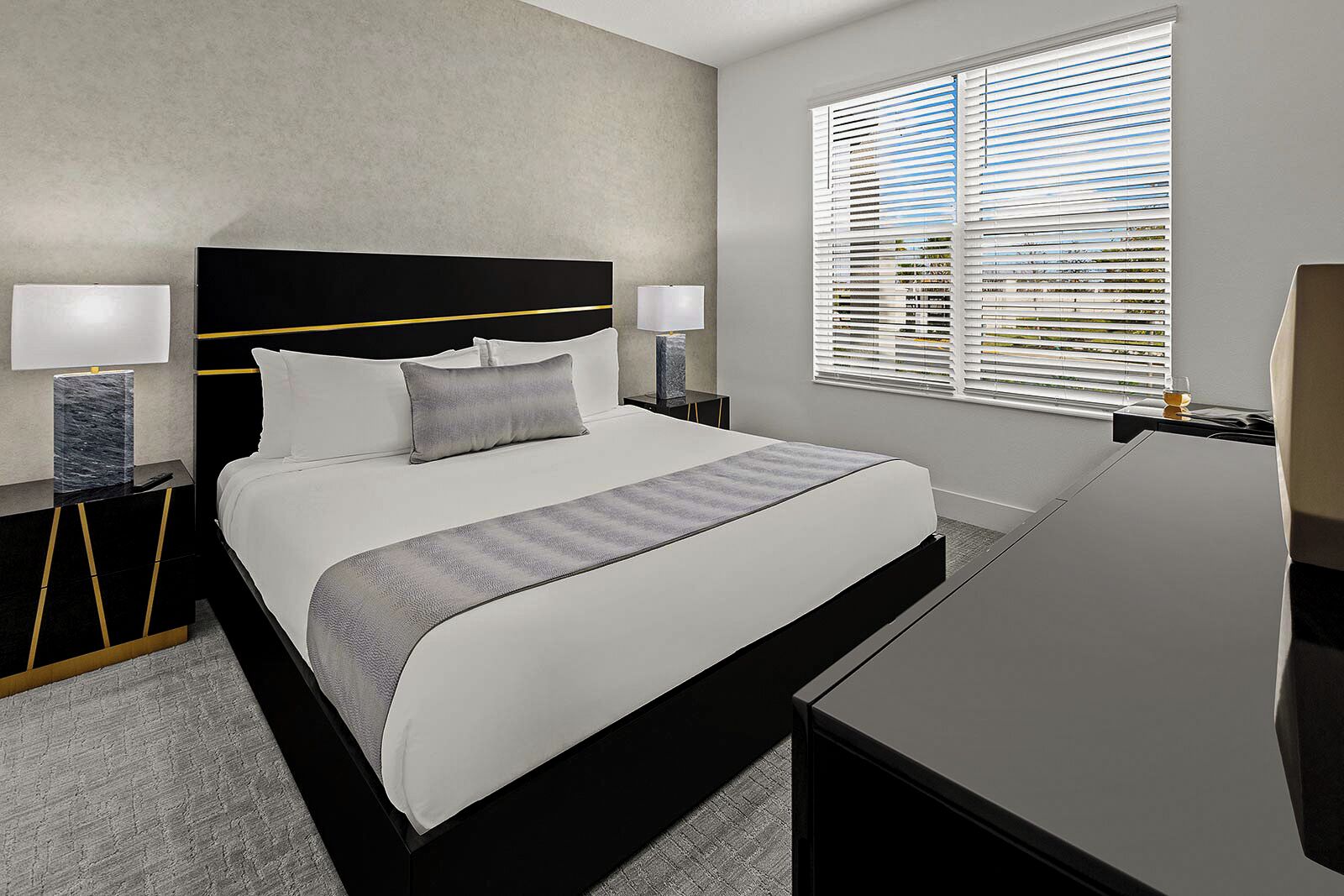 Habitación de hotel moderna con una cama grande, dos mesitas de noche con lámparas, un escritorio y una ventana con persianas.