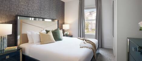 A hotel room with a large bed, a ceiling fan, a nightstand with a lamp, and a view of the outside through a window.