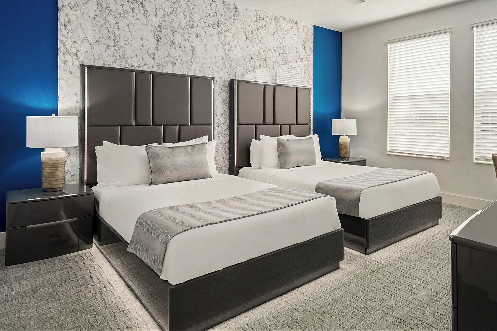 A modern hotel room with two beds, marble-patterned walls, and a large window with blinds.