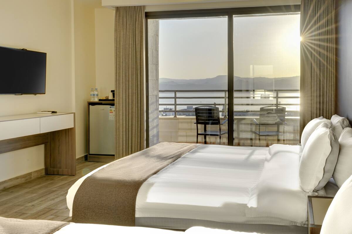 Une chambre d’hôtel équipée d’un lit, d’une télévision, d’un bureau, d’un réfrigérateur et d’un balcon offrant une vue sur la ville et les montagnes.