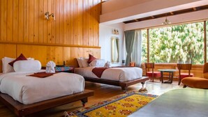 Een hotelkamer met twee bedden, een houten plafond, een groot raam met uitzicht op bomen en een kleine tafel met stoelen.