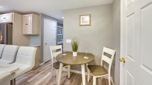 A dining area with a round table, white chairs, and a potted plant. There is a white door and a framed picture on the wall.