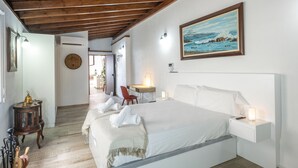 Quarto com teto de madeira, cama com roupa de cama branca, escrivaninha com cadeira e um quadro na parede.