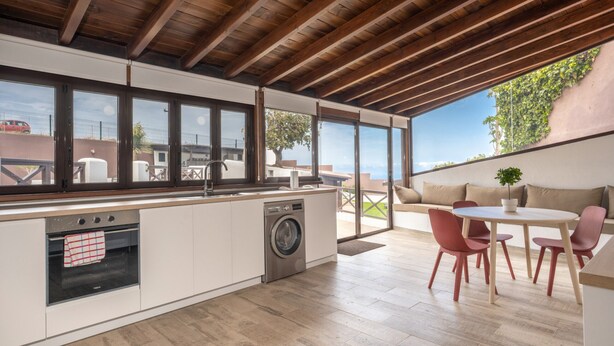Cozinha moderna com forno embutido e máquina de lavar, janelas amplas e área de jantar com cadeiras vermelhas.