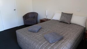 Ein ordentlich bezogenes Bett mit einer grau karierten Tagesdecke, zwei Kissen und zwei gefalteten Handtüchern. Ein grauer Sessel und ein kleiner Nachttisch mit Lampe stehen daneben.