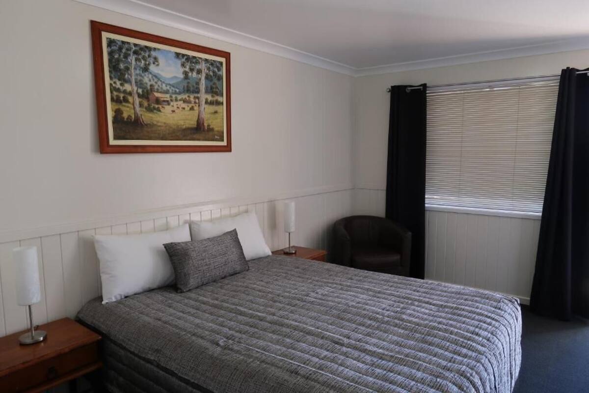 A neatly made bed with a grey quilt, a bedside lamp, and a framed landscape painting on the wall.