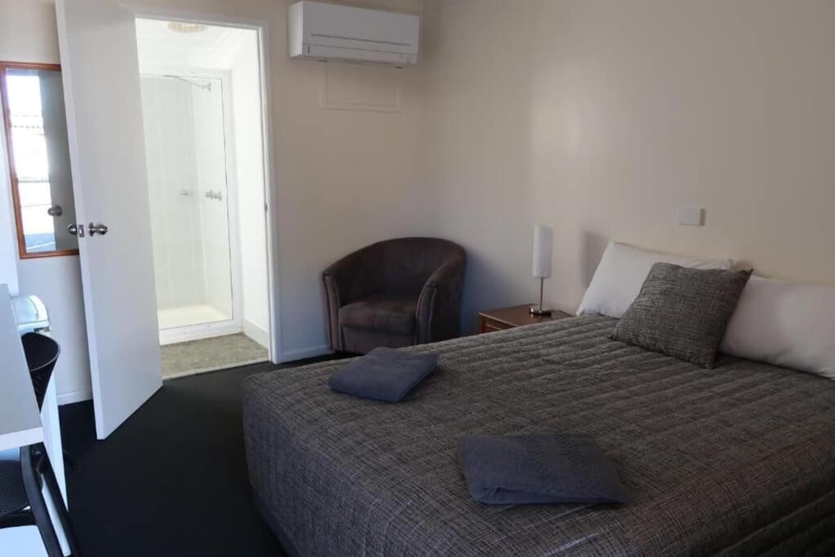 A hotel room with a bed, a chair, a bedside table, and an air conditioning unit.