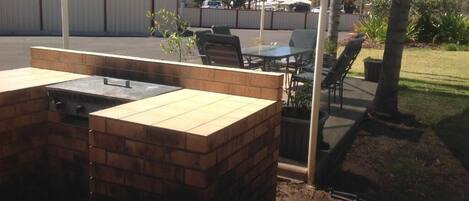 An outdoor patio area with a brick barbecue station, a table, and chairs under a striped umbrella.
