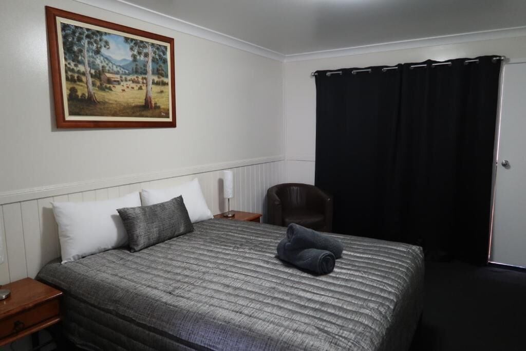 Un lit bien fait, recouvert d’une couvre-lit à rayures, un tableau accroché au mur et un rideau noir à la fenêtre.