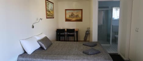 A bedroom with a bed, bedside table, and a desk with chairs. There is a painting on the wall and a door leading to another room.