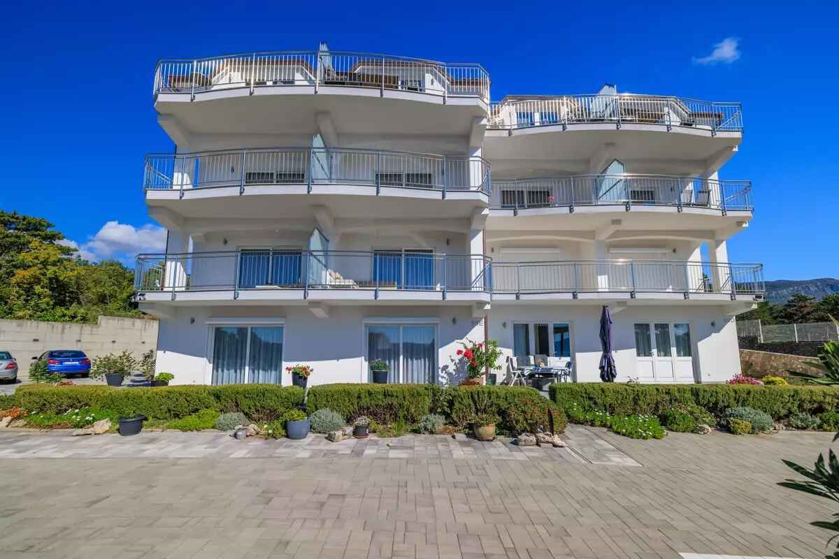 Un edificio residencial de varios pisos con balcones, un camino pavimentado y jardines ajardinados.
