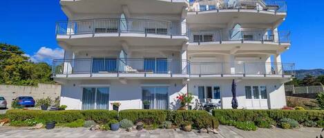 Un edificio residencial de varios pisos con balcones, un camino pavimentado y jardines ajardinados.