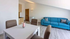 Ein modernes Wohnzimmer mit einem blauen Sofa, einem weißen Esstisch mit Stühlen und einer Küchenzeile im Hintergrund.