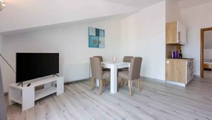 Ein modernes Wohnzimmer mit einem Flachbildfernseher auf einem weißen Schrank, einem Esstisch mit vier Stühlen und einer Küchenzeile mit Holzschränken.