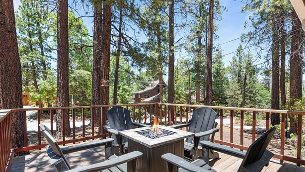 A deck with a square table and four Adirondack chairs, surrounded by tall pine trees.