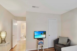 A hotel room with a television on a stand, a chair with a pillow, a bedside table with a lamp, and a door leading to the bathroom.