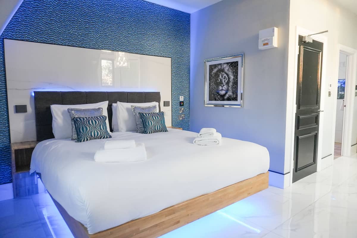 A modern bedroom with a large bed, a headboard with built-in lighting, a framed artwork on the wall, and a visible door to the right.