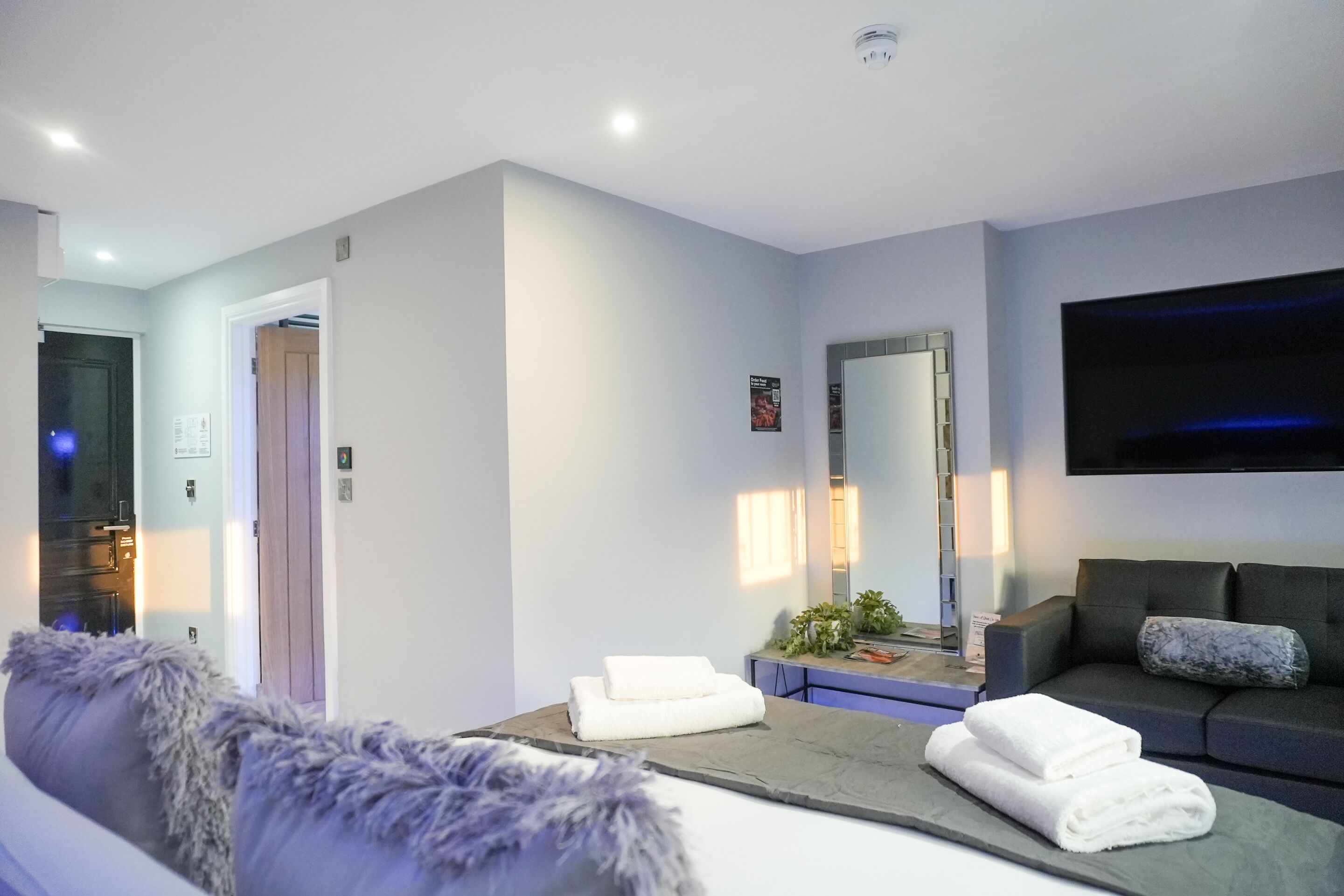 A modern hotel room with a grey sofa, a flat-screen TV, a large mirror, and neatly folded towels on a table.